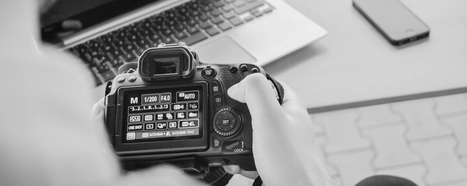 A camera showing its settings screen, held beside a laptop, in black and white